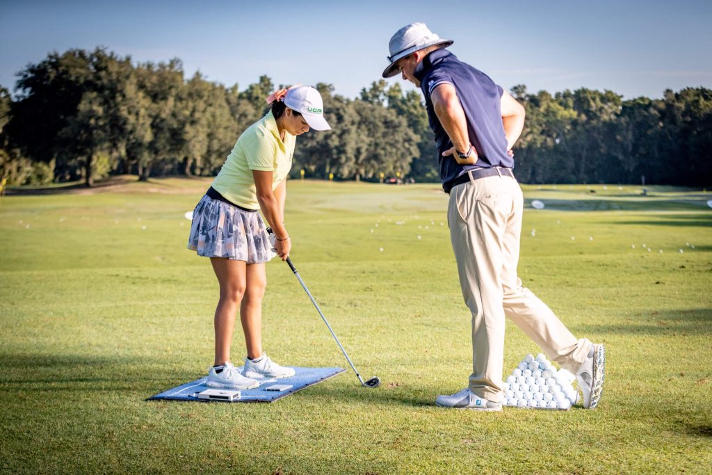 A junior IJGA golfer and coach stand together on the course while the golfer prepares to swing
