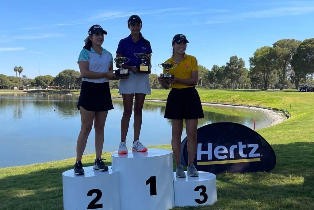 Three female golfers stand on a winner's podium at a golf tournament in Mexico. The podium is tiered by 1st, 2nd, and 3rd place.