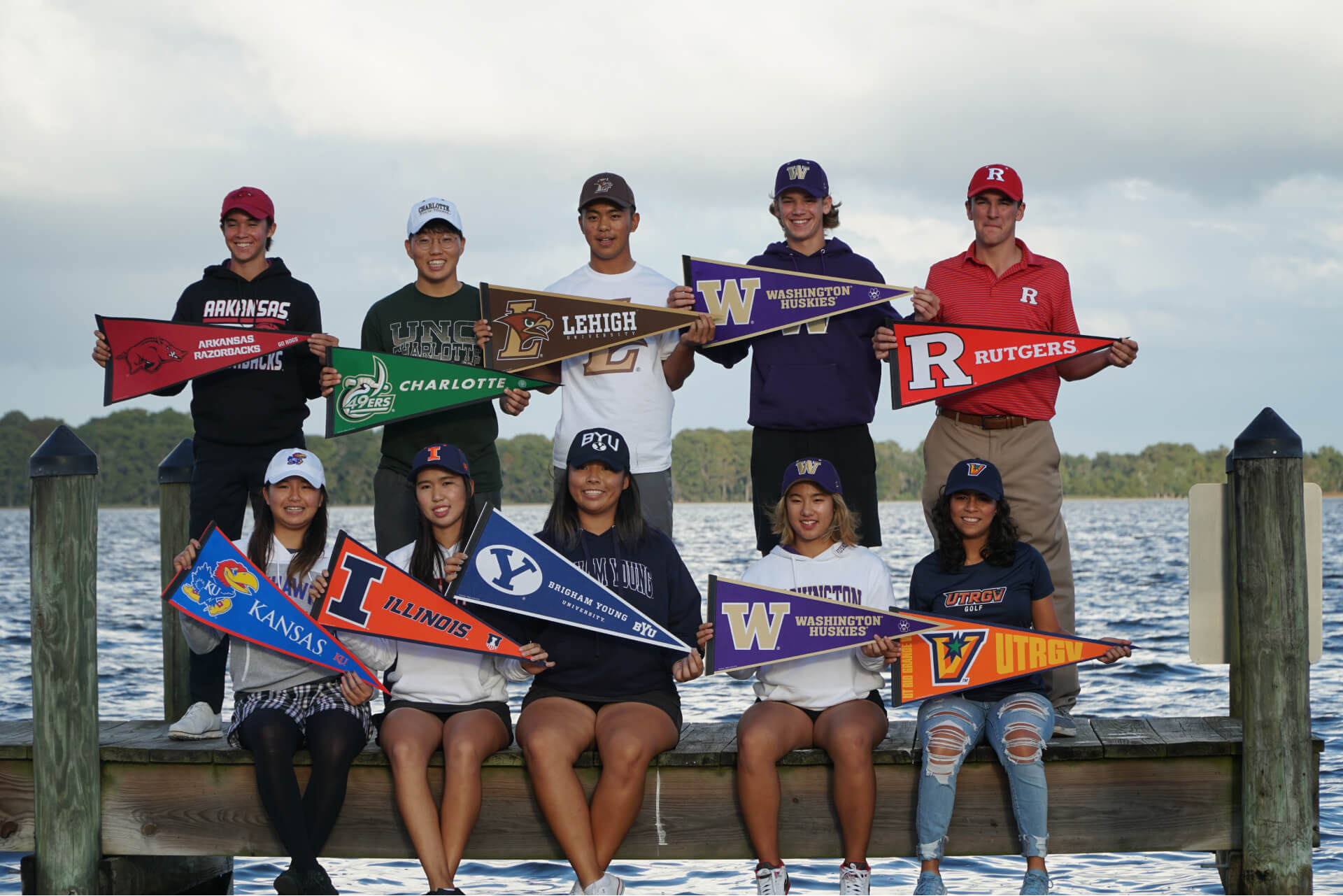 Group of junior-athletes stand and sit together on a dock smiling and holding college pennants