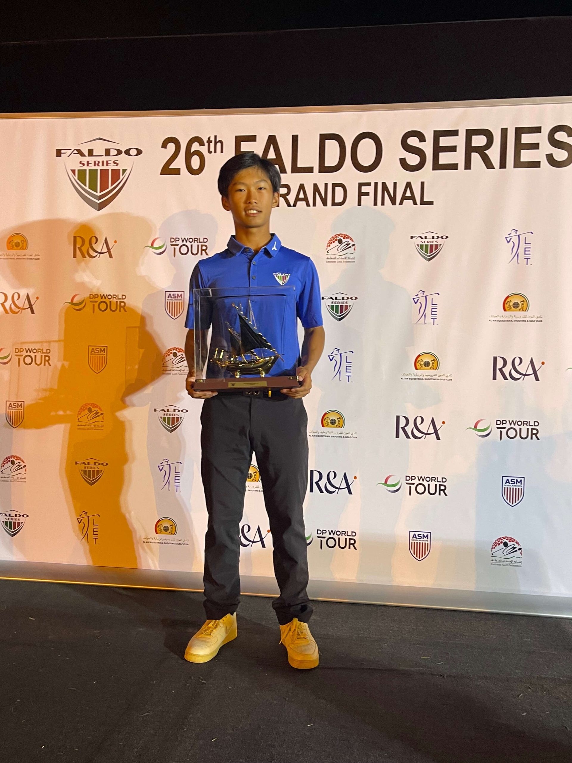 A male golf student stands in front of signage from the Faldo Series Grand Finale. In his hands he is holding a trophy in the shape of a sailboat.