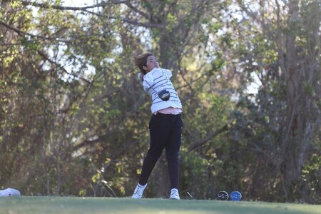 A junior golfer tees off on a tree-lined fairway, captured mid-swing during a tournament round.